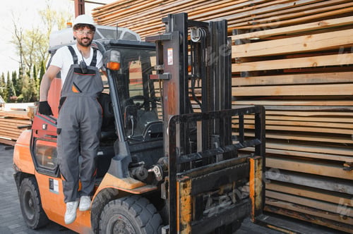Preview: Warehouse worker standing on forklift near lumber stacks