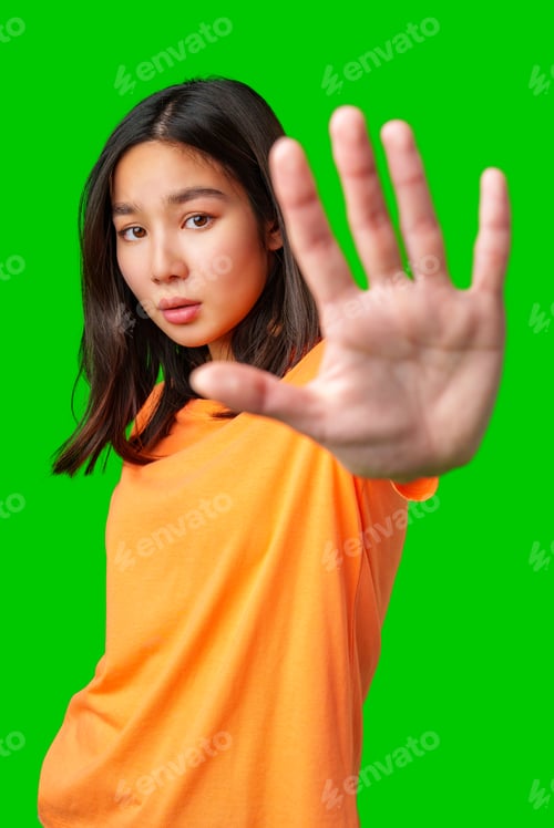 Preview: Young woman in orange shirt poses with hand raised against bright green background