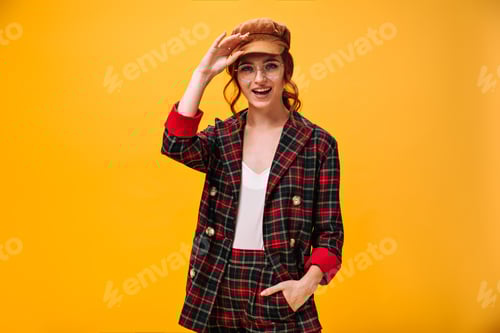 Preview: Lady in cap and coat smiling on orange background. Modern woman with red hair in round glasses and