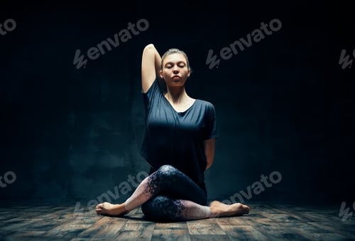 Preview: Young woman practicing yoga doing Gomukhasana, cow face pose, in dark room.