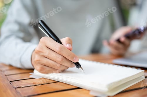 Preview: Close up of a hand holding pen writing in notebook and other hand holding smartphone. Study outdoor.