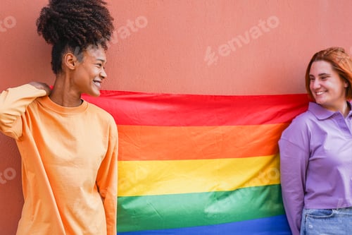 Preview: Happy female gay couple having fun together at LGBT pride event - Lesbian girls holding rainbow flag