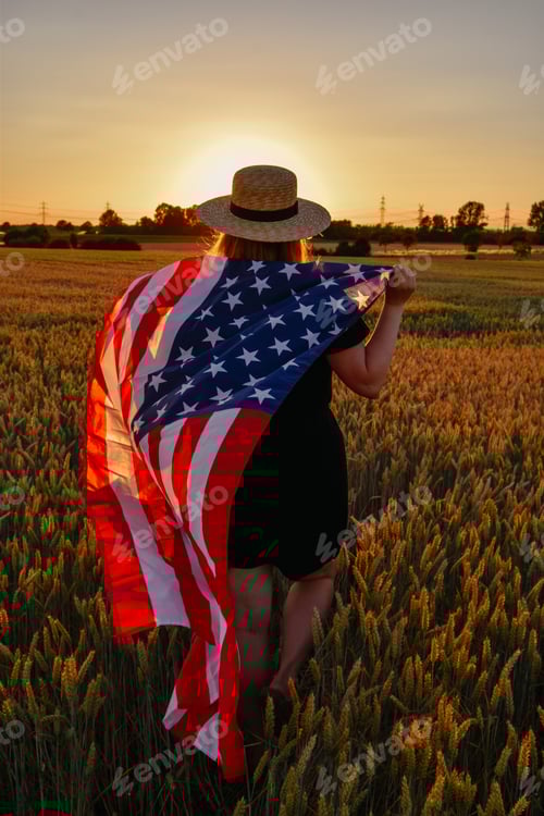 Preview: Woman walking with flag of USA in cornfield at sunset