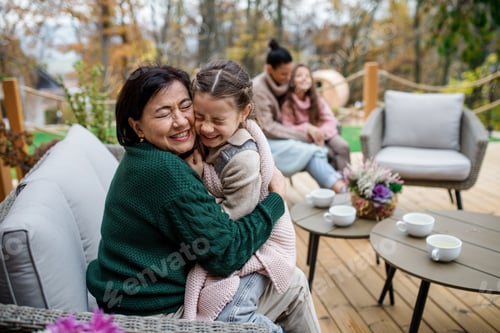Preview: Happy little girl with grandmother sitting wrapped in blanket outdoors in patio in autumn