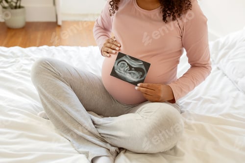 Preview: Unrecognizable black pregnant woman showing baby sonography photo while sitting on bed