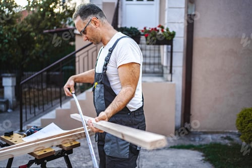 Preview: Mature male carpenter measuring wooden plank outdoors