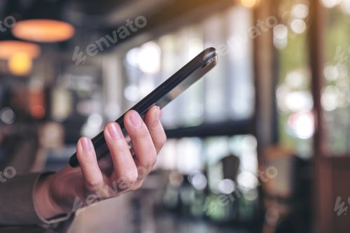 Preview: A woman holding and using a black smart phone with blur background in cafe