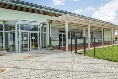 Vorschau: Außenansicht einer modernen Schule mit gemähten Grasfeldern und gepflastertem Boden, der dorthin führt