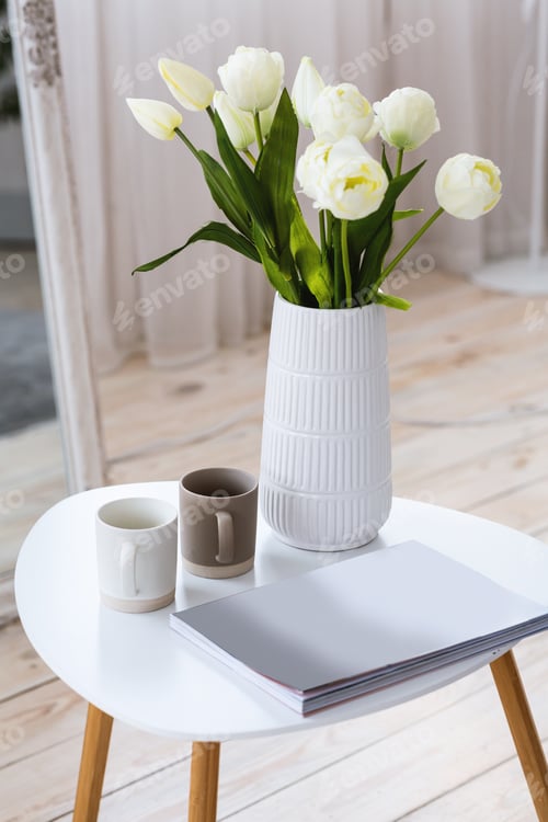 Preview: White tulips in vase on table with book or notebook and two cups on table on wooden floor