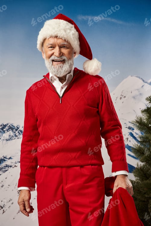 Preview: joyous Santa in warm red outfit posing with gift bag with mountain backdrop, winter concept