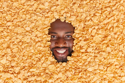 Preview: Positive dark skinned man with thick beard buried in dry cornflakes smiles pleasantly going to prepa