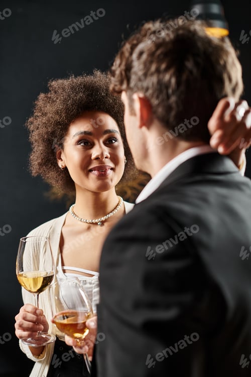 Preview: happy african american woman holding wine glass and looking at man during date on valentines day