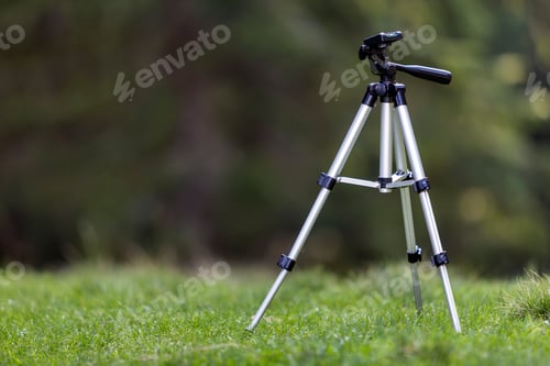 Preview: Isolated aluminum shiny tripod standing alone in fresh grassy meadow on bright sunny summer day on
