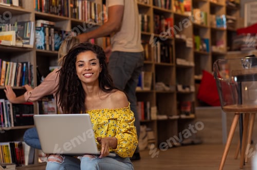 Preview: Portrait of young female student sitting at library using laptop with her friends in background.