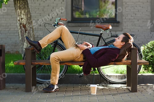 Preview: smiling young freelancer lying on bench and using laptop