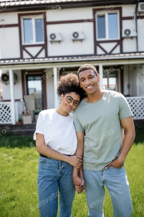 Preview: smiling african american couple holding hands and looking at camera near new house