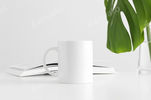 Preview: White mug mockup with a monstera leaf in a vase and a book on a white table.
