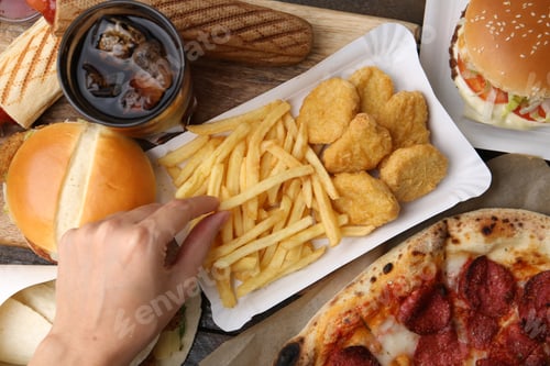 Preview: Woman taking tasty French fry at wooden table with different fast food and cola, top view