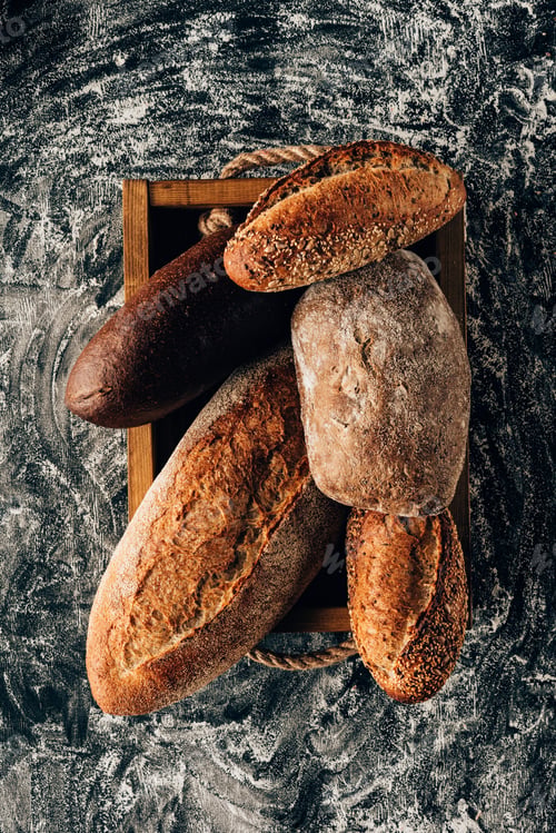 Preview: top view of arranged loafs of bread in wooden box on dark tabletop with flour