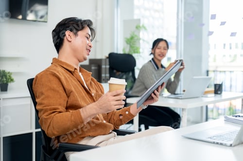 Preview: A positive Asian male office worker reading a document while enjoying a hot drink at his desk.