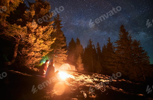 Preview: Pair of vacationers sitting near campfire outdoors under stars.