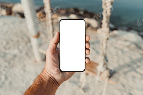Preview: Man standing by the sea and holding mobile smart phone with blank mockup screen, closeup of hand