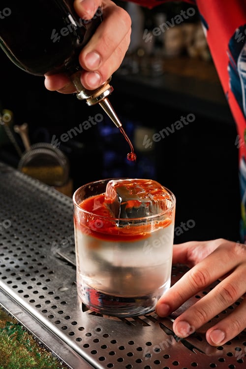 Preview: Bartender adding bitter to a transparent cocktail in a rocks glass with a crystal clear ice cube