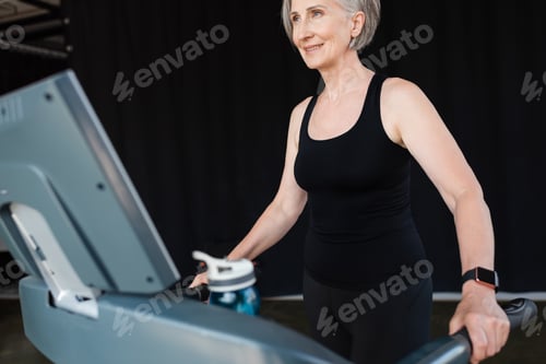 Preview: positive senior woman with grey hair working out on treadmill in gym