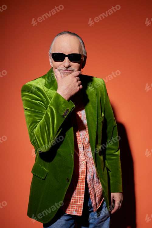 Preview: smiling senior man in dark sunglasses and green velour blazer touching beard and looking at camera