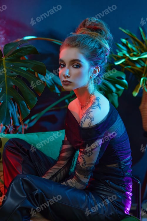 Preview: Young woman, wearing a black sheer dress, poses with tropical plants in front of a blue and red neon