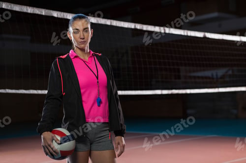 Preview: Mid-adult female coach holding volleyball with whistle lanyard in sportswear on gym court with net