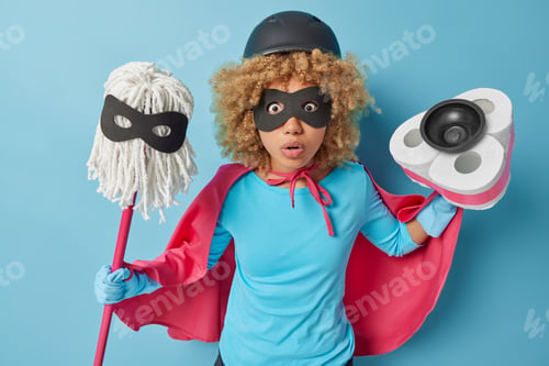 Preview: Frightened shocked curly haired woman dressed in superhero costume holds tools for cleaning prepares