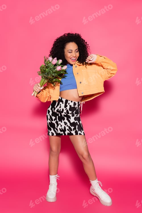 Preview: full length of happy african american woman holding bouquet of flowers on pink