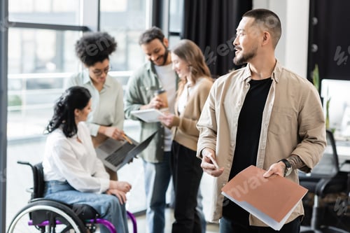 Preview: cheerful asian man holding smartphone and folder near multicultural team on blurred background