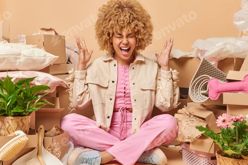 Preview: Stressed curly haired woman gestures angrily screams loudly sits crossed legs surrounded by