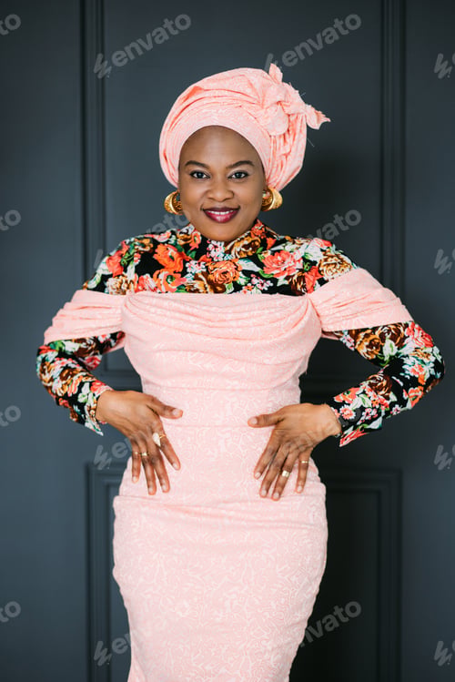 Preview: Portrait of charming African woman wearing stylish pink dress and turban on head, smiling on camera