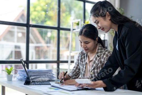 Preview: Two women are sitting at a desk, one of them writing on a piece of paper