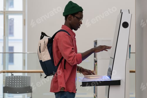 Preview: Young African student guy standing in library using self-service terminal to borrow or return books
