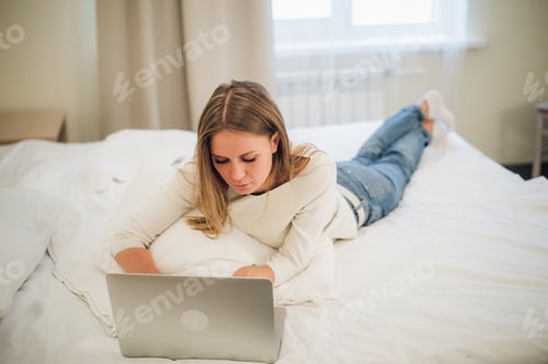 Vista previa: Una linda jovencita sonriendo emocionada ante la pantalla de su portátil. Se está relajando en la cama.