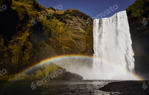 Preview: Stunning view of Skogafoss waterfall in Iceland with a vibrant rainbow arching across the mist.