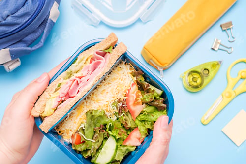 Preview: Female hands holding plastic lunch box with sandwich and salad on blue background top view.
