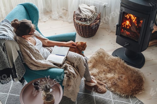 Preview: Young woman write in a notebook sitting in a armchair by the fi