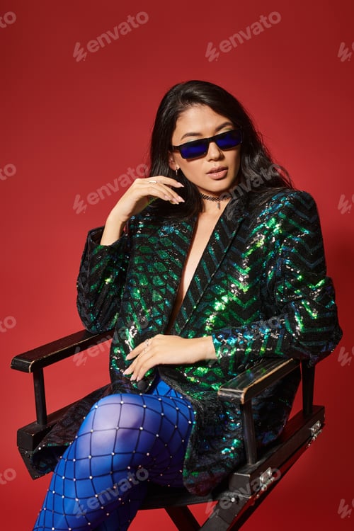 Preview: pretty asian woman in sunglasses and green jacket with sequins sitting on chair on red backdrop