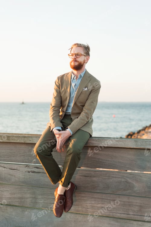 Preview: Full length of businessman sitting on retaining wall against beach