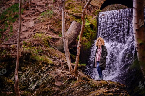 Preview: Young woman in green parka stands above waterfall in mountains