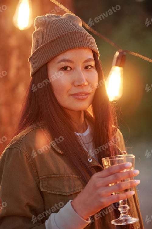 Preview: Happy Asian girl with dark long hair toasting with drink against lights