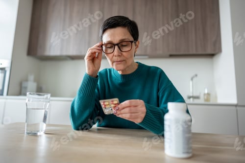 Preview: Worried mature woman adjusts glasses reading medication blister pack before taking pills at home