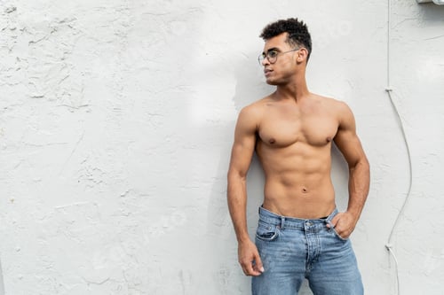 Preview: cuban man with athletic body posing in blue jeans and trendy round-shaped eyeglasses near building