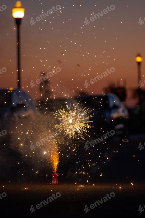 Preview: Fireworks sparkle as the sun sets. Warm orange and gold colors with street lights in the background.