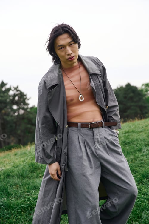 Preview: Stylish young asian man poses in a gray trench coat in a serene park setting